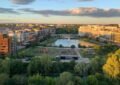 Vue aérienne en plongée du parc du Port-Marianne à Montpellier, montrant un étang central avec une fontaine, des pelouses vertes, des immeubles modernes environnants et des arbres sous un ciel clair de fin d'après-midi.