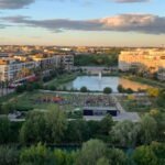 Vue aérienne en plongée du parc du Port-Marianne à Montpellier, montrant un étang central avec une fontaine, des pelouses vertes, des immeubles modernes environnants et des arbres sous un ciel clair de fin d'après-midi.