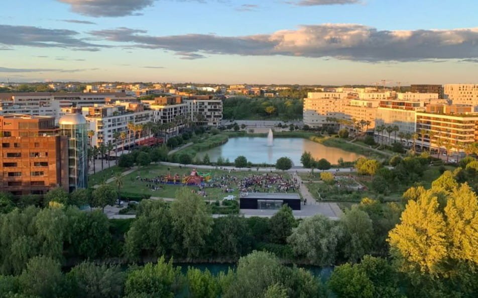 Vue aérienne en plongée du parc du Port-Marianne à Montpellier, montrant un étang central avec une fontaine, des pelouses vertes, des immeubles modernes environnants et des arbres sous un ciel clair de fin d'après-midi.