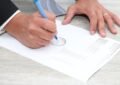 Main droite écrivant sur un document avec un stylo bleu, main gauche posée dessus, table en bois