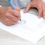 Main droite écrivant sur un document avec un stylo bleu, main gauche posée dessus, table en bois